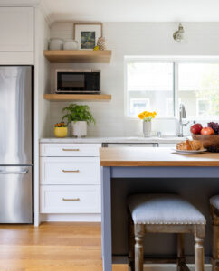 Kitchen with open wood shelves, white drawer cabinets with brass pulls, stainless refrigerator and blue island stools