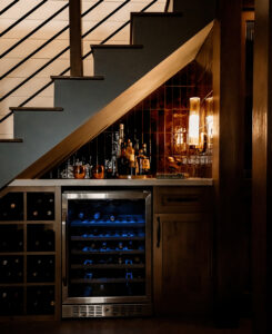 Detail of a lit wine and bar nook under the stairs, with glossy dark tile backsplash and a wine refrigerator