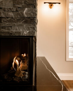 Detail of a gas fireplace framed by dark stacked stone, with a black stone-look ledge and wall sconce