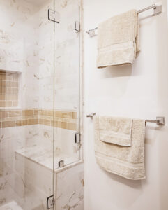 Glass shower enclosure with a marble-look tile bench and tan accent border, beside two towel bars