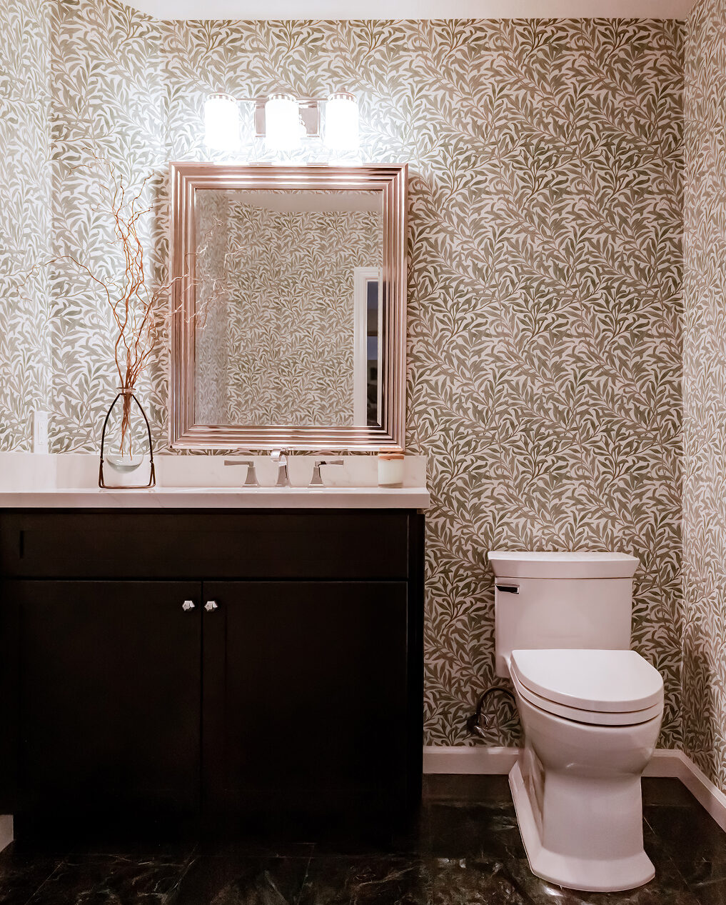 Powder room with leaf-pattern wallpaper, a dark shaker-style vanity, a three-light fixture and a white toilet