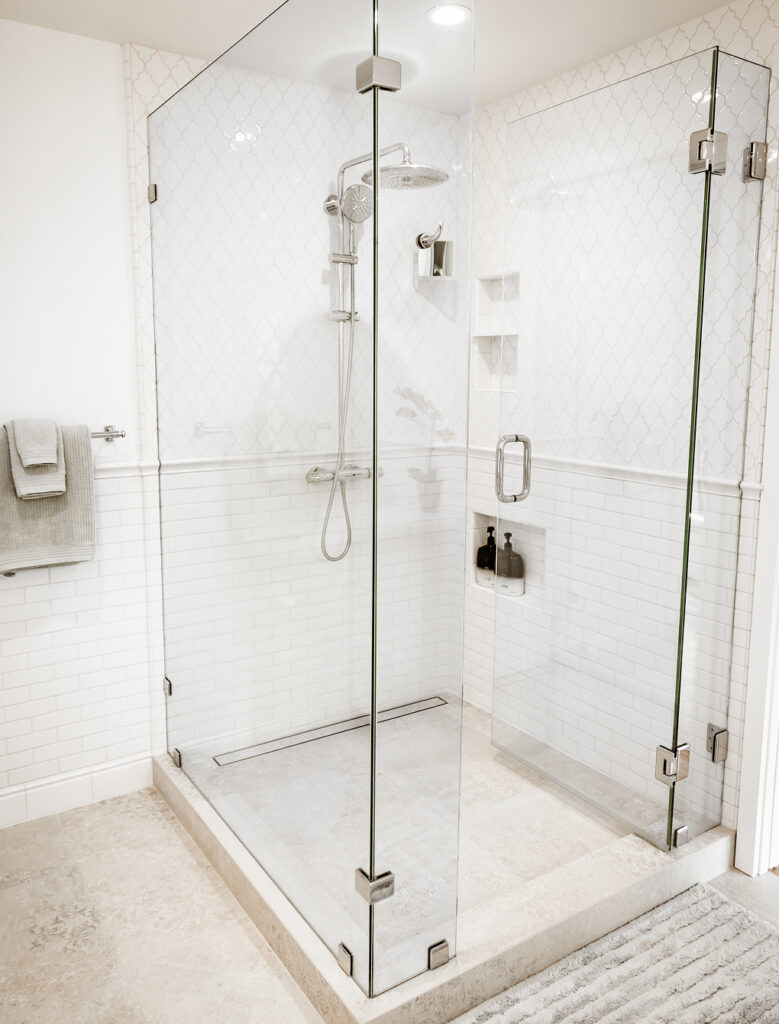 Corner glass shower with white arabesque and subway tile, a rain showerhead, wall niches and a linear floor drain
