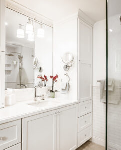 Vanity with white shaker cabinets, a tall storage cabinet, three-light sconce over a large mirror and a white countertop