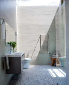 Primary bathroom seen from the doorway, with a tall stone-look tile wall, towel warmer and pebble floor in sunlight