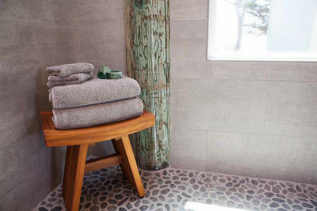 Wood stool with folded gray towels beside a glass-paneled shower with a pebble tile floor