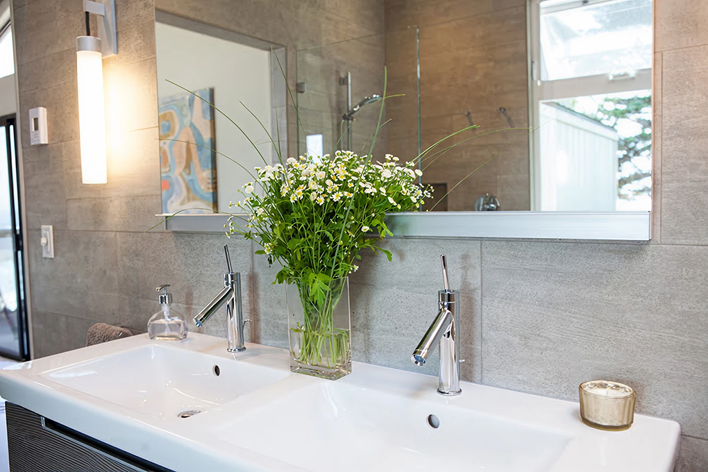Double-sink vanity detail with two chrome faucets, a wide mirror and a wall sconce