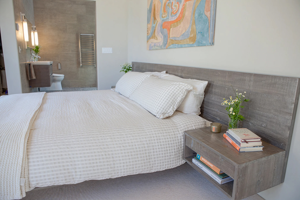 Bed with a weathered wood headboard and floating nightstand, with the bathroom vanity in the background