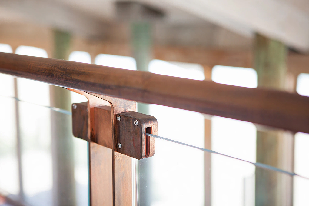 Close-up of a loft railing with a wood handrail, copper-toned brackets and glass panels